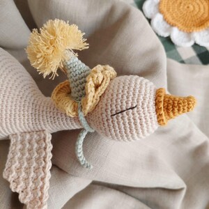 May include: A handmade crochet goose doll in cream, yellow, and light blue. The goose wears a party hat with a yellow pom-pom and a yellow collar. The doll is lying on a beige linen surface, with a daisy coaster in the background.