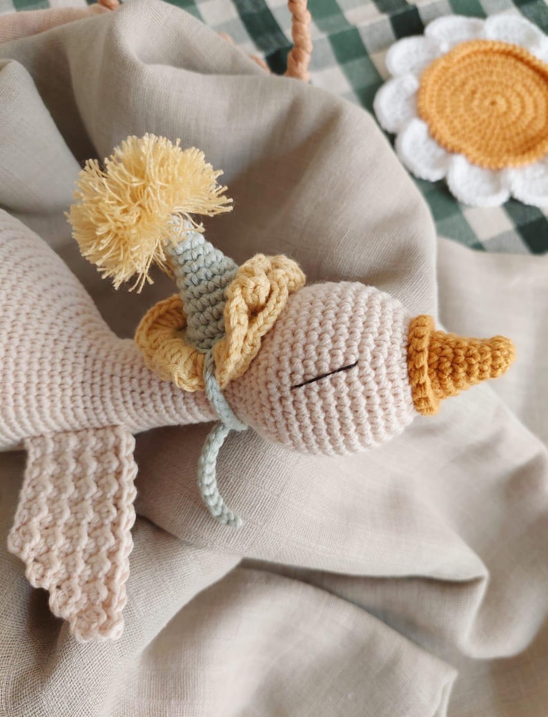 May include: A handmade crochet goose doll in cream, yellow, and light blue. The goose wears a party hat with a yellow pom-pom and a yellow collar. The doll is lying on a beige linen surface, with a daisy coaster in the background.