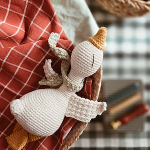 May include: A handmade crochet goose doll with a yellow hat and feet, and a green scarf, resting in a woven basket. The goose is cream-colored, with detailed stitching. It rests on a red and white checkered cloth.