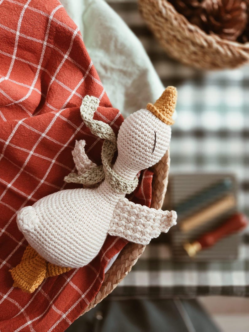 May include: A handmade crochet goose doll with a yellow hat and feet, and a green scarf, resting in a woven basket. The goose is cream-colored, with detailed stitching. It rests on a red and white checkered cloth.