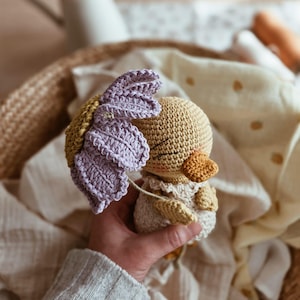 Pode incluir: Um brinquedo de patinho de crochê feito à mão com um acessório em forma de flor. O patinho é principalmente bege com um bico laranja e um vestido branco. A flor é lavanda e amarela. O brinquedo é segurado em uma cesta de vime.