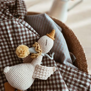 May include: A handmade crochet goose toy with a party hat and a yellow pom-pom. The goose is light beige with yellow and blue accents. It rests in a woven basket with brown and white checkered fabric and blue striped fabric.