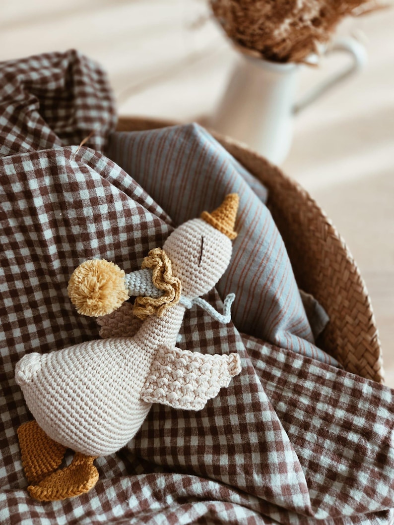 May include: A handmade crochet goose toy with a party hat and a yellow pom-pom. The goose is light beige with yellow and blue accents. It rests in a woven basket with brown and white checkered fabric and blue striped fabric.