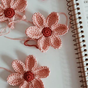 May include: Close-up of several handmade crochet flowers. The flowers have pink petals and a dark red centre. Some have loose yarn attached. The flowers are on a white surface, near a spiral notebook.