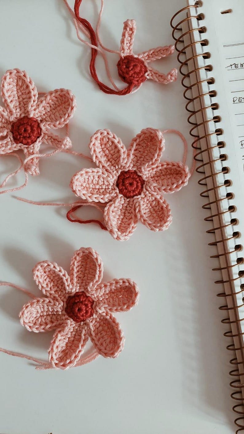 May include: Close-up of several handmade crochet flowers. The flowers have pink petals and a dark red centre. Some have loose yarn attached. The flowers are on a white surface, near a spiral notebook.