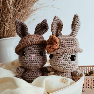 Pode incluir: Duas bonecas de coelho de crochê feitas à mão em uma cesta branca. Os coelhos são cinza e marrons, usam chapéus e têm olhos pretos e narizes rosados. A cesta é forrada com um tecido creme. Plantas secas estão no fundo.