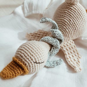 May include: A handmade crochet duck toy in beige, light blue, and mustard yellow. The duck has a rounded body, a long neck, and a detailed beak. The wings are beige and the duck is lying on a white cloth.