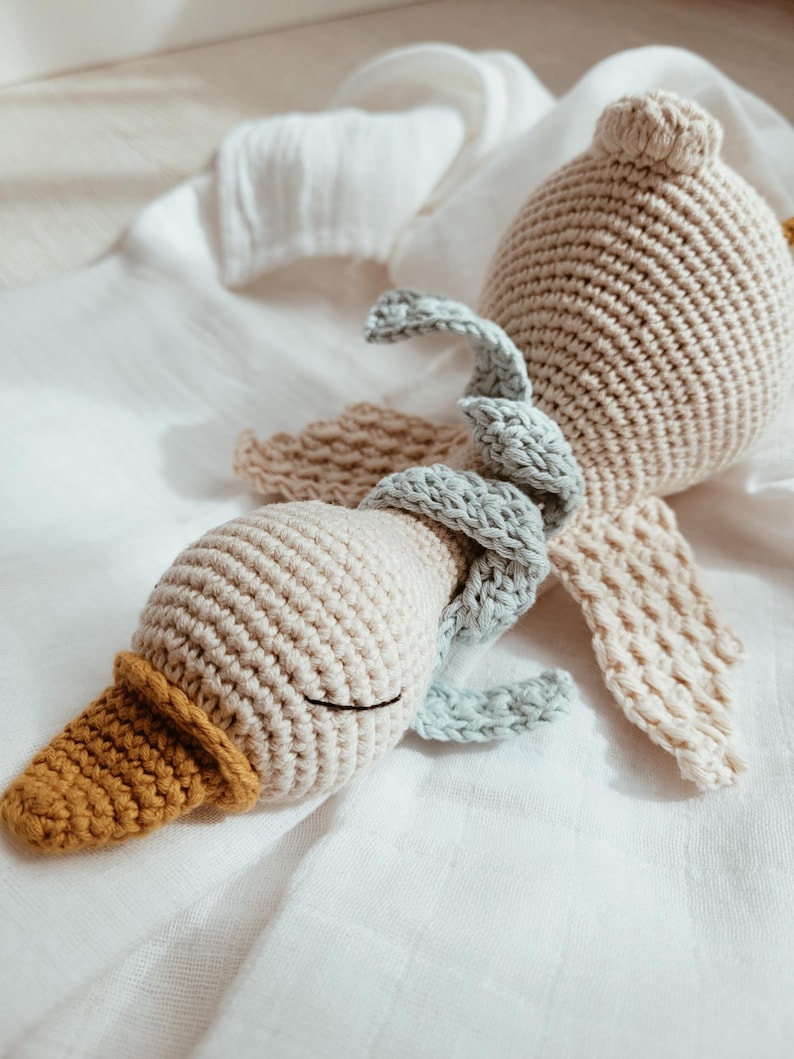 May include: A handmade crochet duck toy in beige, light blue, and mustard yellow. The duck has a rounded body, a long neck, and a detailed beak. The wings are beige and the duck is lying on a white cloth.