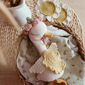 May include: A white crocheted goose toy featuring a pink floral crown, a yellow scalloped cape, and orange feet. The goose has closed eyes and is nestled inside a woven basket lined with a light green fabric featuring a small floral pattern.