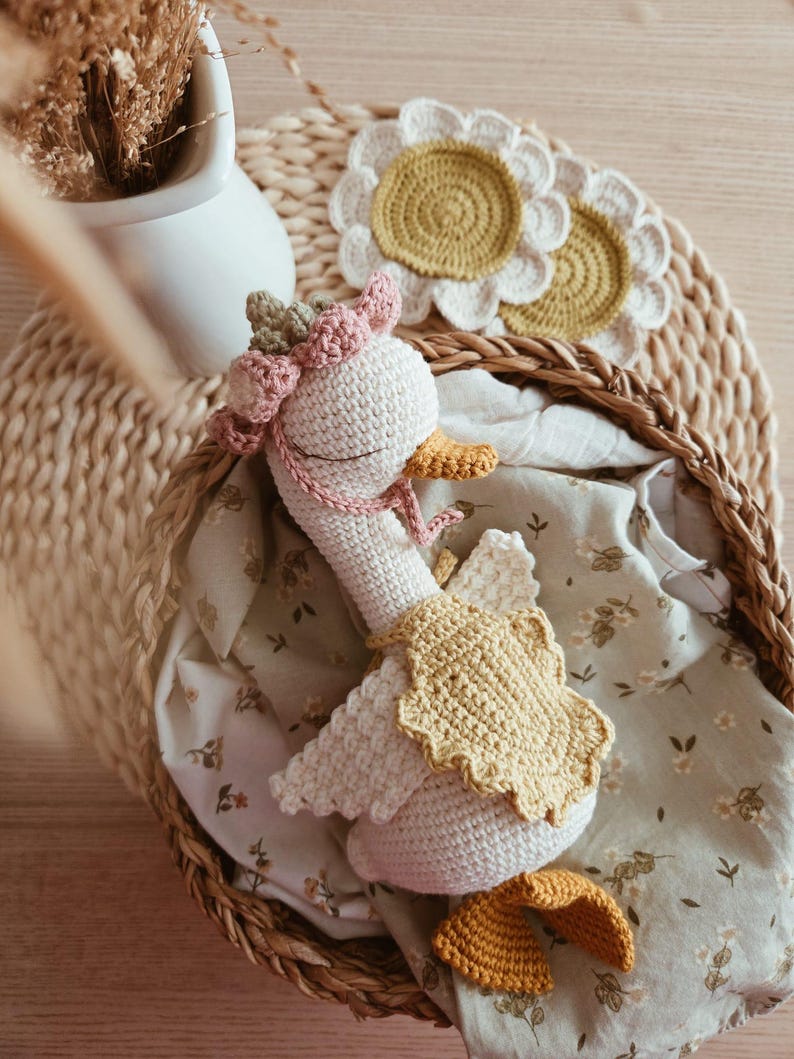 May include: A white crocheted goose toy featuring a pink floral crown, a yellow scalloped cape, and orange feet. The goose has closed eyes and is nestled inside a woven basket lined with a light green fabric featuring a small floral pattern.