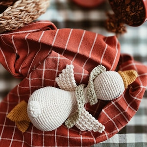 May include: A handmade, white crochet swan doll with green and yellow accents, resting on a red and white checkered cloth. The swan has a long neck and detailed wings. The background includes a woven basket and a red container.