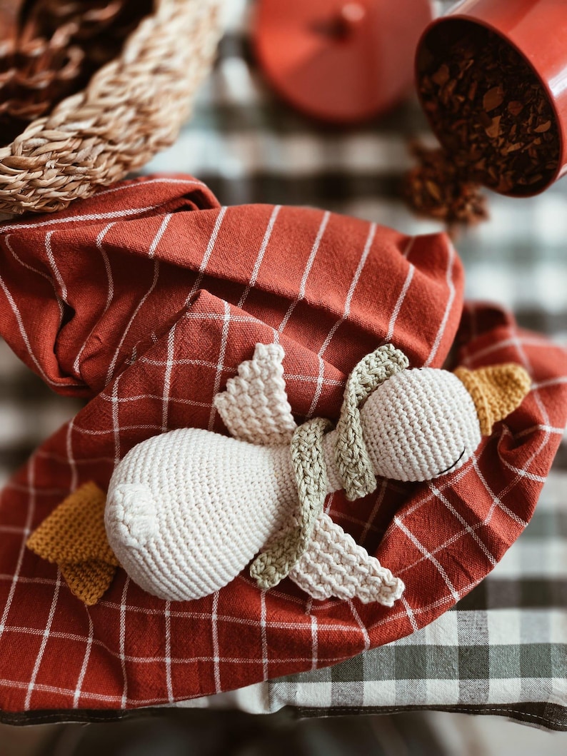 May include: A handmade, white crochet swan doll with green and yellow accents, resting on a red and white checkered cloth. The swan has a long neck and detailed wings. The background includes a woven basket and a red container.