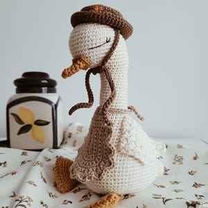 May include: A crochet goose doll featuring a cream-colored body, a brown hat with a small orange detail, and a brown apron. The goose has a yellow beak, orange feet, and embroidered closed eyes. It is crafted with visible yarn stitches.