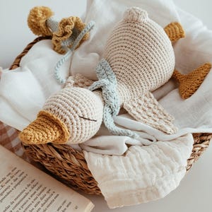 May include: A handmade crochet duck toy in a woven basket. The duck is beige with a yellow beak and feet, and a light blue scarf. The basket also contains a white cloth and a crochet flower. A charming, soft toy.