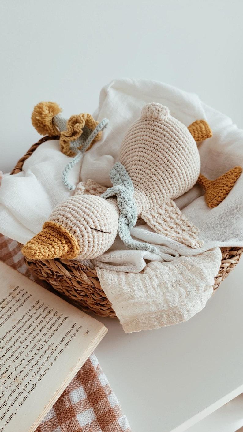 May include: A handmade crochet duck toy in a woven basket. The duck is beige with a yellow beak and feet, and a light blue scarf. The basket also contains a white cloth and a crochet flower. A charming, soft toy.