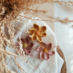 May include: Three handmade crochet flowers in shades of yellow and pink rest on a white cloth. The flowers have a detailed petal design and a contrasting centre. The image is a close-up, with dried grasses in the background.