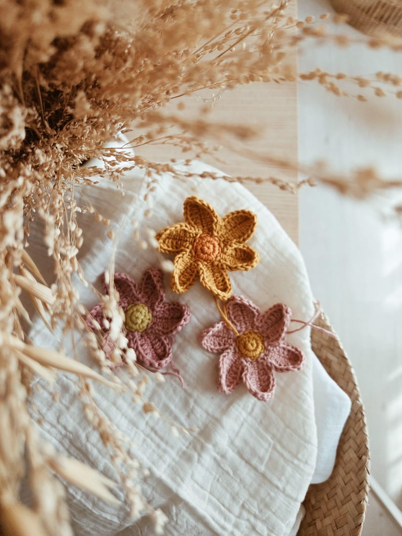 May include: Three handmade crochet flowers in shades of yellow and pink rest on a white cloth. The flowers have a detailed petal design and a contrasting centre. The image is a close-up, with dried grasses in the background.