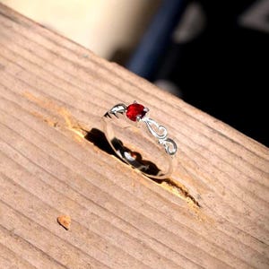 Puede incluir: Un anillo de plata con una piedra preciosa roja redonda y facetada engastada en el centro. La banda está elaborada con un delicado diseño de volutas caladas que se extiende desde el engaste a ambos lados, creando un patrón metálico.