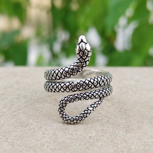 Puede incluir: Un anillo de metal plateado con forma de serpiente enroscada. El diseño presenta un patrón de escamas texturizado y detallado en todo el cuerpo de la serpiente, con la cabeza elevada en la parte superior y la cola curvada hacia el interior.