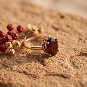 Puede incluir: Un anillo de oro con una gran piedra preciosa de color rojo intenso y corte ovalado, engastada en una cesta de metal decorativa. El anillo está colocado junto a un pequeño racimo de bayas rojas secas y diminutas flores secas claras.