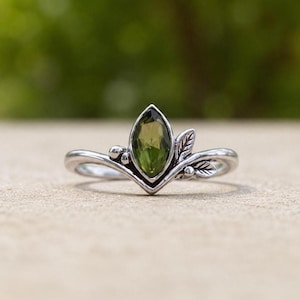 Puede incluir: Un anillo de plata con una piedra preciosa de color verde oliva en talla marquesa en el centro. La banda tiene forma de V y está decorada con pequeños motivos de hojas metálicas y diminutas esferas a ambos lados de la piedra central.