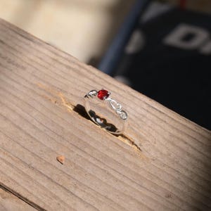 May include: A silver ring featuring a small, round, vibrant red gemstone set in the centre. The band is crafted with delicate, openwork scroll patterns that extend from the setting along both sides of the polished metallic ring shank.