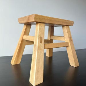 May include: A small, light-coloured wooden stool with a rectangular seat and four angled legs. The stool is constructed with visible joinery and has a natural wood grain finish. The stool is approximately 30 cm tall.