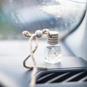 Puede incluir: Un pequeño ambientador para coche de cristal transparente con tapón de madera y cuerda decorativa. El frasco tiene forma geométrica y una etiqueta con las letras "R M". La cuerda es beige con detalles amarillos.