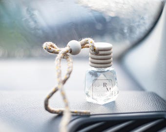 May include: A small, clear glass car air freshener with a wooden cap and a decorative rope. The bottle has a geometric shape and a label with the letters "R M". The rope is beige with yellow accents.
