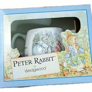 Könnte beinhalten: Eine Peter Rabbit Tasse von Wedgwood in einer hellblauen Box. Die Tasse zeigt Illustrationen von Peter Rabbit und anderen Charakteren. Die Box hat ein Fenster, das die Tasse zeigt.