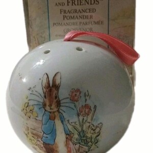 May include: A white ceramic spherical pomander featuring a printed illustration of Peter Rabbit wearing a blue jacket, surrounded by flowers. The object has small ventilation holes at the top and a pink ribbon loop for hanging, with a box labelled Peter Rabbit and Friends Fragranced Pomander behind it.