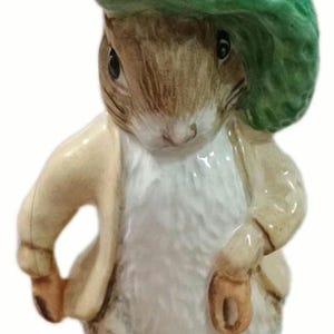 Puede incluir: Figura de cerámica de un conejo con sombrero verde y chaqueta beige. El conejo tiene pelaje marrón y está de pie sobre un trozo de hierba verde. El sombrero tiene un pequeño acento naranja.