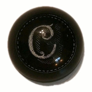 May include: A black, round shift knob with a stitched border. The center features a white, stylized letter "C". The knob has a glossy finish and is set against a white background.