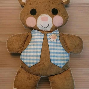 Puede incluir: Un peluche de oso pardo con un chaleco a cuadros azul y blanco y una flor. El oso tiene ojos negros, mejillas rosas y una boca sonriente. Las orejas y los pies tienen detalles rosas.