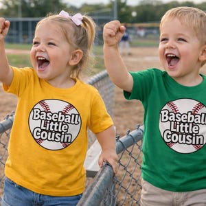 May include: Two young children wearing yellow and green t-shirts with a baseball graphic that says "Baseball Little Cousin". They are smiling and raising their fists in the air, near a chain-link fence.