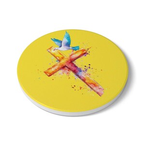 Può includere: Un sottobicchiere rotondo e giallo con un design ad acquerello. Il design presenta una croce nei toni dell'arancione, del rosso e del viola, con una colomba blu che vola sopra. Schizzi aggiungono all'aspetto artistico. Il sottobicchiere ha un bordo bianco.