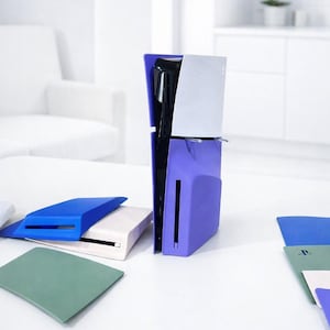 May include: A purple and silver game console with matching colored faceplates and accessories. The console is standing upright on a white table, with additional faceplates in blue, white, and green.