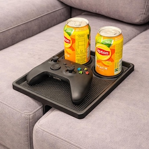 Könnte beinhalten: Ein schwarzes Sofatablett mit strukturierter Oberfläche, das zwei Dosen Lipton Pfirsich-Tee und einen schwarzen Videospiel-Controller hält. Das Tablett ist für die Montage an einer Sofalehne konzipiert.