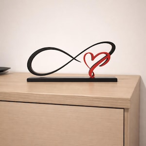 May include: A black infinity symbol sculpture with a red heart detail. The sculpture is mounted on a rectangular black base, placed on a light wood surface. The design is a modern representation of love and eternity.