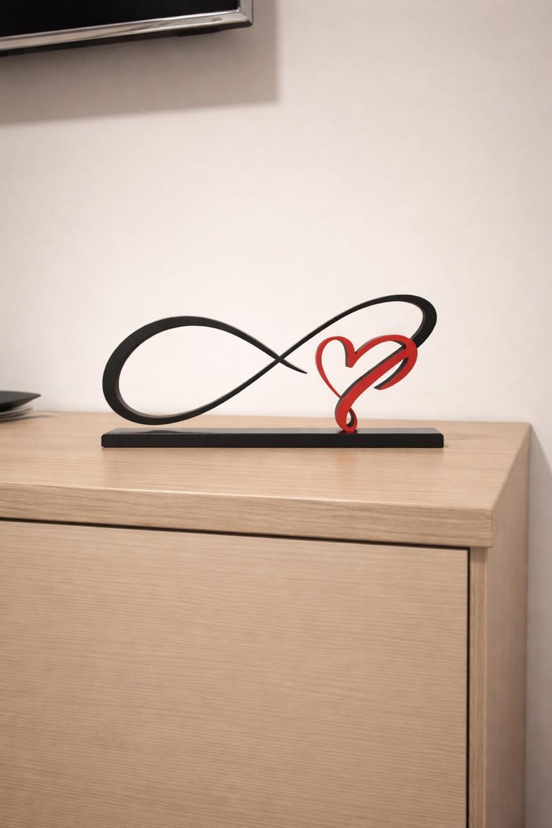 May include: A black infinity symbol sculpture with a red heart detail. The sculpture is mounted on a rectangular black base, placed on a light wood surface. The design is a modern representation of love and eternity.
