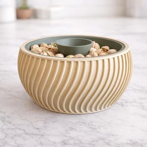 Puede incluir: Un cuenco de servir beige con un diseño ondulado, interior verde oscuro y un cuenco para salsa central más pequeño. El cuenco está lleno de pistachos, perfecto para aperitivos.