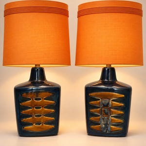 May include: A pair of table lamps featuring dark blue ceramic bases with a central vertical panel of textured, golden-brown geometric cutouts. Each lamp is topped with a cylindrical orange fabric lampshade with horizontal ribbed detailing.