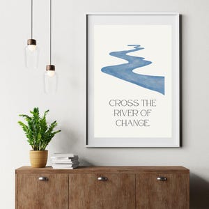 Puede incluir: Un cuadro enmarcado con un diseño de río azul sobre un fondo crema. El texto "CROSS THE RIVER OF CHANGE" está impreso debajo del río. La obra de arte se exhibe sobre un mueble de madera.