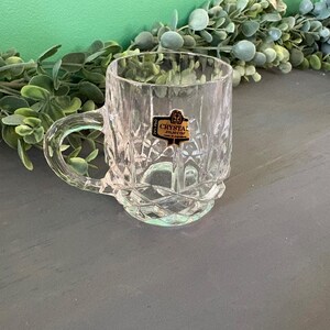 May include: A clear crystal mug with a handle, featuring a diamond-cut pattern. The mug has a sticker that reads "CRYSTAL zajecar". The mug is sitting on a gray surface with green foliage in the background.