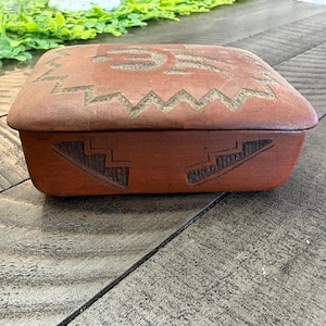 May include: A rectangular, reddish-brown ceramic box with a lid. The lid features a carved design with a central motif and a zigzag border. The sides of the box have carved geometric patterns. The box is likely used for storage.