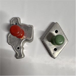 May include: Two metal cookie cutters with wooden handles. One cutter is shaped like the state of Texas with a red handle, and the other is a diamond shape with a green handle. Both cutters feature small circular holes in the metal frame.