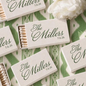 May include: Off-white rectangular matchboxes featuring the dark green printed text The Millers and the date 7.6.26 in a cursive script. Each box is partially open, revealing a row of wooden matches with white tips inside the cardboard sleeve.