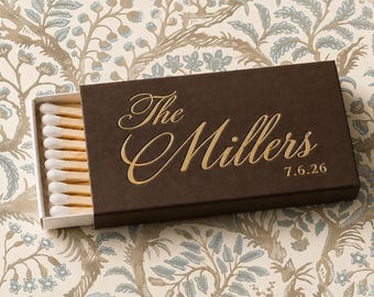 May include: A rectangular dark brown matchbox with a sliding tray containing wooden matches with white tips. The top of the box features gold foil lettering that reads The Millers and the date 7.6.26 in a cursive and sans-serif font style.