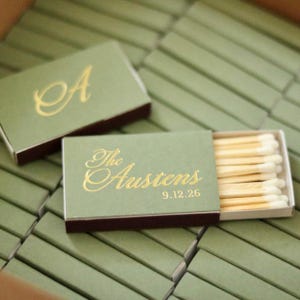 May include: A collection of rectangular matchboxes with sage green covers and dark brown edges. One box is open, revealing wooden matches with white tips. Gold foil text on the boxes reads "A" or "The Austens" with the date "9.12.26".