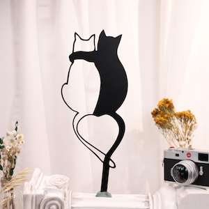 Könnte beinhalten: Schwarze Metall-Katzen-Silhouette, die eine weiße herzförmige Form umarmt. Die Skulptur steht auf einem grünen Sockel. Eine Vintage-Kamera und Trockenblumen befinden sich im Hintergrund.